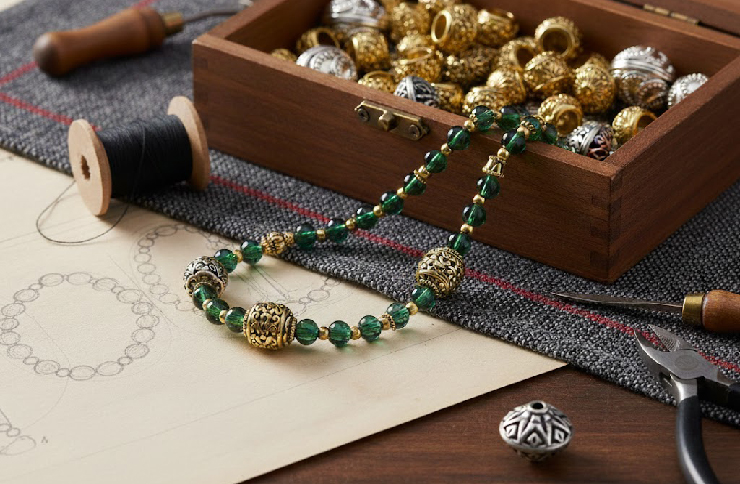 Detailed Balinese brass beads and silver accents displayed in a wooden craft box next to a handmade green crystal necklace and jewelry tools.